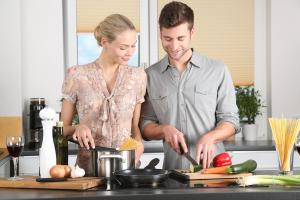 Eine Frau und ein Mann stehen in der Küche und kochen. Die Frau steht am Kochtopf und rührt Nudeln in den Topf, der Mann schneidet eine Zucchini.