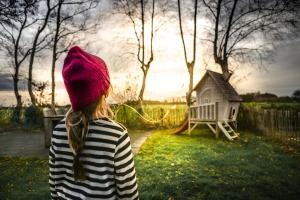 ein Mädchen mit einer roten Mütze steht im Garten und blickt in die Ferne. Im Hintergrund ist ein Gartenhaus, Bäume und der Sonnenuntergang zu sehen.
