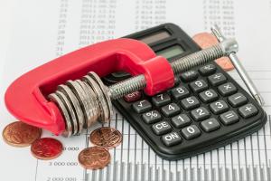 Eine rote Schraubzwinge mit Geld liegt auf einem Taschenrechner, um Sparmaßnahmen aufzuzeigen