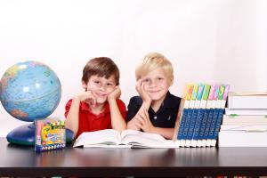 zwei Kinder Sitzen an einem Tisch, vor ihnen liegt ein aufgeschlagenes Buch, auf der rechten Seite steht ein Globus, auf der linken Seite verschiedene Bücher
