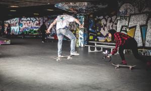 ein Jugendlicher macht vor einer Graffitiwand einen Sprung auf seinem Skatboard. Ein zweiter Skaterboarder filmt ihm dabei