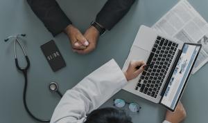 Eine Ärztin mit Stift in der Hand sitzt am Laptop und berät einen ihr gegenübersitzenden Patienten. Außerdem liegt vor Ihr eine Brille, ein Smartphone und ein Stethoskop.