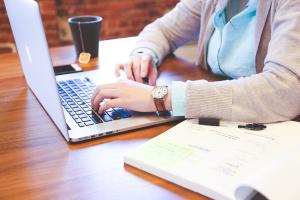 Eine Frau sitzt am Laptop und recherchiert. Rechts neben ihr liegen ein Notizbuch und ein Stift.