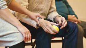 zwei Hände eines älteren Menschens sind im Mittelpunkt des Bildes. In der rechten Hand hält er ein Ball. Ein junger Mensch hält mit seiner linken Hand das rechte Handgelek des älteren Menschens.