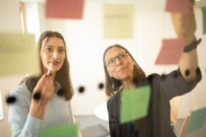 Zwei Frauen sind druch eine transparente Oberfläche zu sehen, an der transparenten Fläche sind Klebezettel und Klebepunkte zu sehen