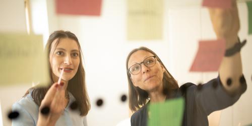 Zwei Frauen sind druch eine transparente Oberfläche zu sehen, an der transparenten Fläche sind Klebezettel und Klebepunkte zu sehen